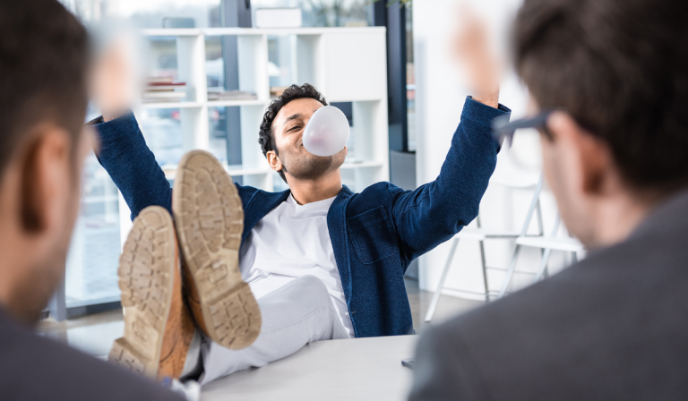 Man with his feet up on a conference table in a meeting, blowing bubblegum and making a rude gesture.